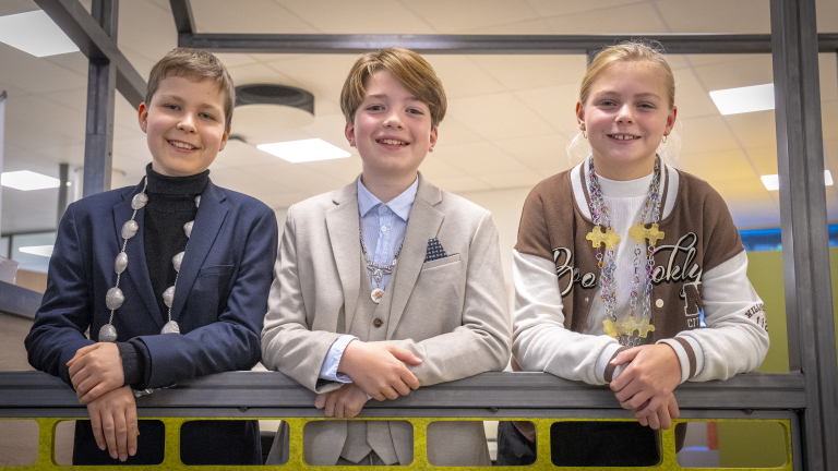 Drie jonge kinderen lachen en dragen een ketting met een medaille, terwijl ze leunen op een hek binnenshuis.