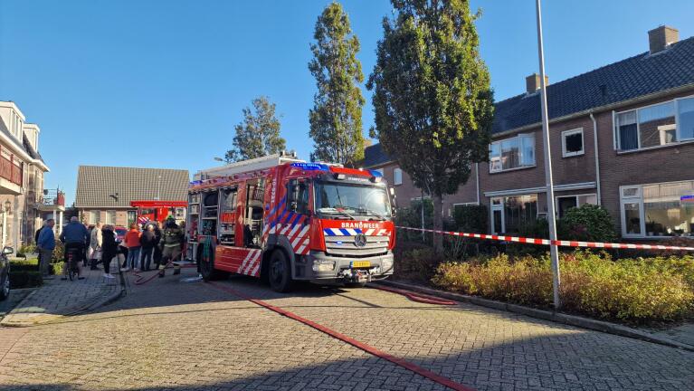 Brandweerwagen geparkeerd in een woonstraat, omringd door toeschouwers en politieafzetting, met een huis op de achtergrond.