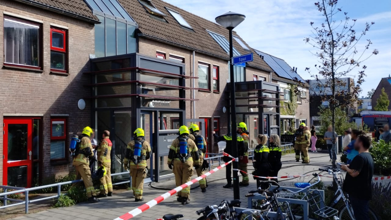 Brandweermannen in actie bij een rijtjeshuis in woonwijk, afgezet met rood-wit lint, met enkele omstanders en politieagenten.