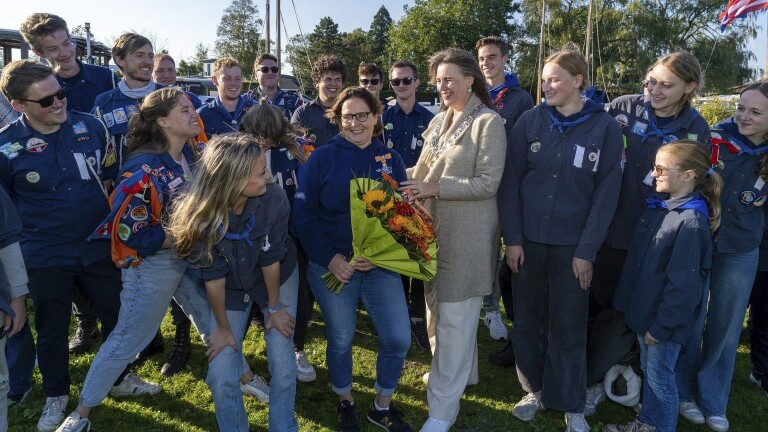 Een groep scouts, gekleed in blauw met insignes, omringt een vrouw die bloemen vasthoudt. Ze staan buiten op een grasveld.