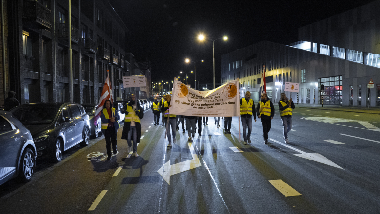 Een groep mensen in gele hesjes loopt 's avonds over straat met een spandoek en protestborden.