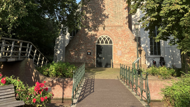 Kerkingang met stenen muur, bruggetje en bloeiende rozenstruiken op de voorgrond.