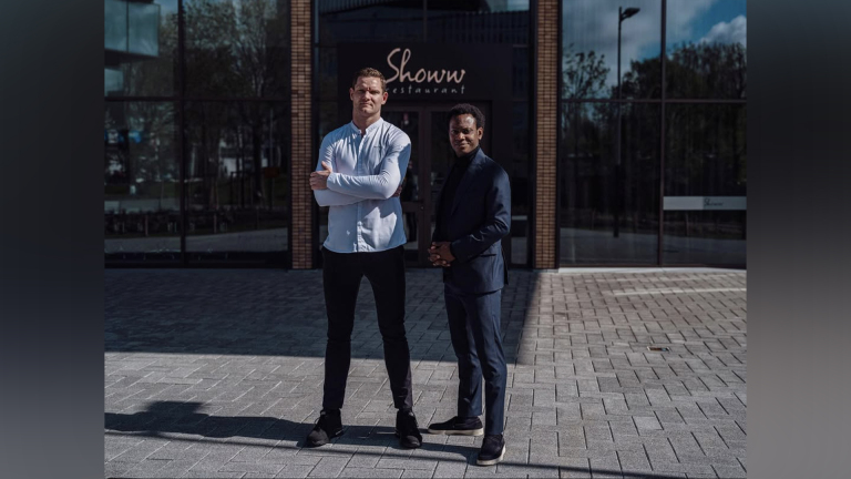 Twee mannen poseren voor een restaurant met de naam "Shouw" op de achtergrond.