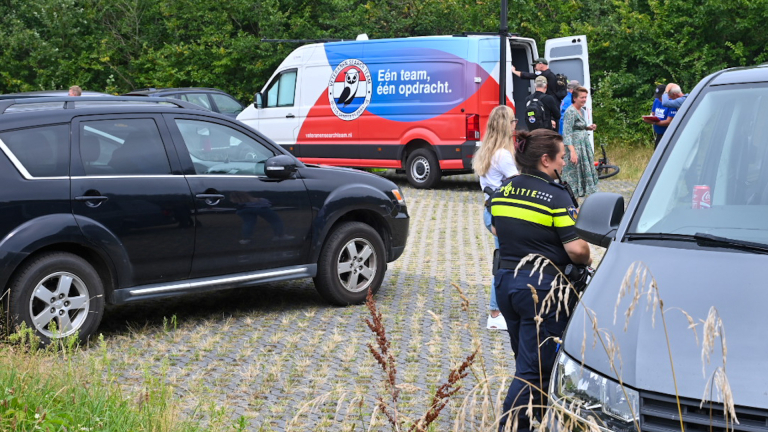 Politieagenten en mensen bij een parkeerplaats met verschillende auto's, waaronder een busje met opschrift "Eén team, één opdracht."