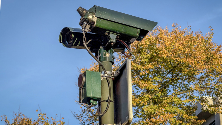 Groene bewakingscamera op een paal, met bomen en blauwe lucht op de achtergrond.