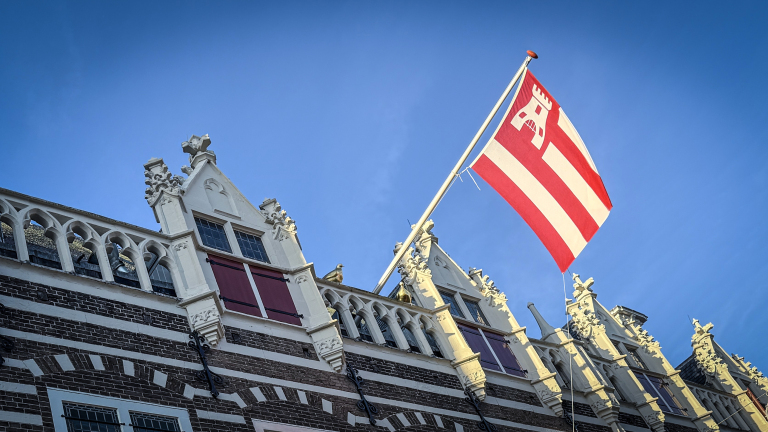 Oude gevel met sierlijke architectuur en een rood-witte vlag, tegen een blauwe lucht.