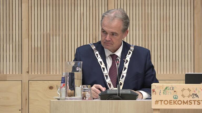 Man in een pak met ambtsketen zit achter een microfoon, met een karaf water en een houten hamer op tafel.