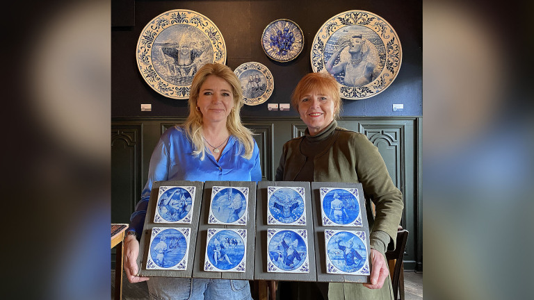 Twee vrouwen poseren met blauwe keramische tegels in een kunstgalerie met grote versierde borden aan de muur.