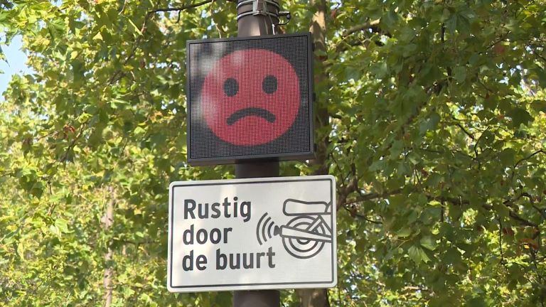 Waarschuwingsbord met een rood, sip gezicht en tekst "Rustig door de buurt" tegen groene bladeren achtergrond.