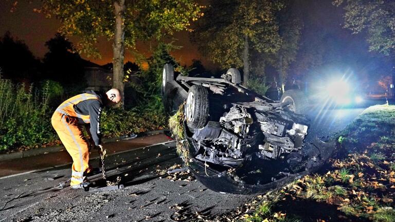 Auto op de kop na een ongeval op een donkere weg, terwijl een persoon in oranje werkkleding de plek onderzoekt met een ketting.