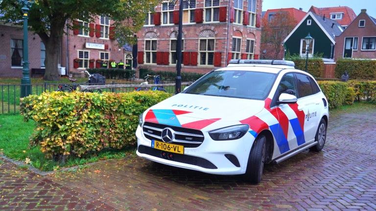 Een Nederlandse politieauto geparkeerd op een straat met een groene heg en een gebouw op de achtergrond.