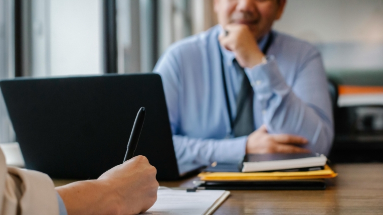 Twee mensen in een gesprek, met een pen op papier en een laptop op tafel.