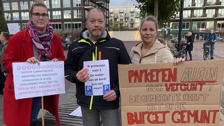 Drie mensen houden protestborden vast tegen parkeerbeleid in een stedelijke omgeving.