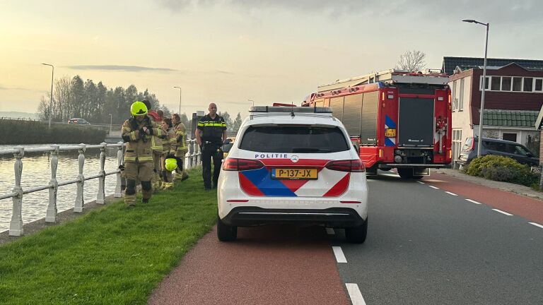 Brandweer- en politievoertuigen geparkeerd langs een weg bij een kanaal; brandweerlieden naast het water en een agent naast een politieauto.