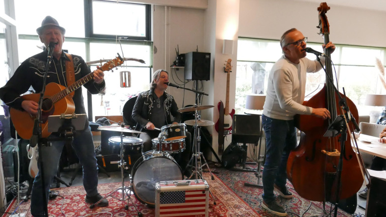 Drie muzikanten spelen muziek; een speelt gitaar, een ander drums, en de derde speelt contrabas. Ze zingen samen in een kamer met tapijt en muziekapparatuur.