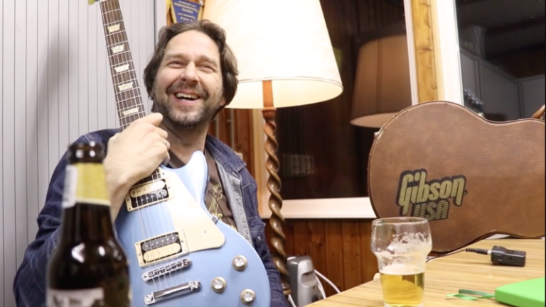 Man lacht terwijl hij een blauwe gitaar vasthoudt, met een biertje en een Gibson gitaarkoffer op de voorgrond.