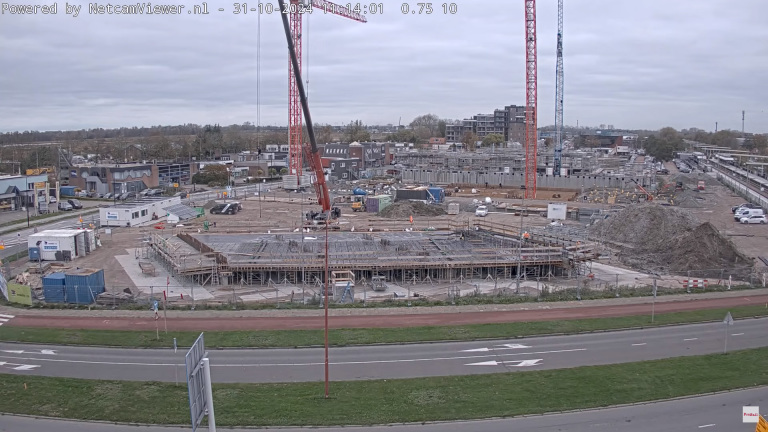 Bouwplaats met hijskranen, betonconstructies en bouwmaterialen, omringd door wegen en gebouwen.