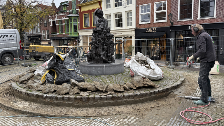 Een man werkt aan een renovatie rond een standbeeld op een plein, omringd door bouwmaterialen en apparatuur. Op de achtergrond zijn winkels en historische gebouwen te zien.