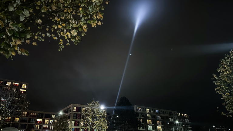 Nachtelijke scène met appartementen en boomtoppen, met een heldere lichtstraal die naar de hemel schijnt.