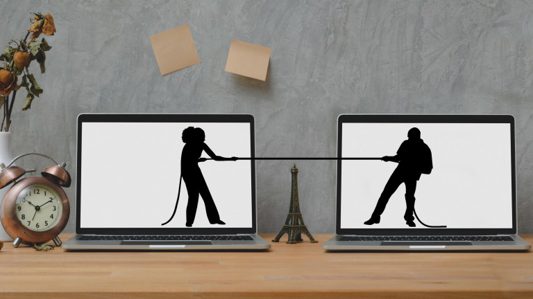 Twee laptops met silhouetten van mensen die een touwtrekken wedstrijd houden, gescheiden door een miniatuur Eiffeltoren op een houten tafel met een wekker en een vaas met verwelkte bloemen.