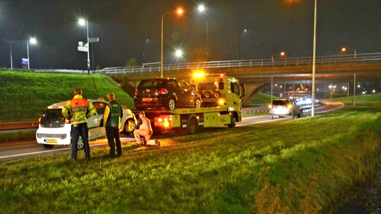 Een bergingsvoertuig met waarschuwingslichten staat stil op de snelweg met een zwarte auto op de laadbak; mensen staan naast een witte auto ernaast.