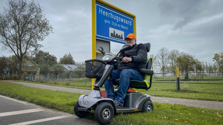 Man op scootmobiel bij bord "Heerhugowaard, gem. Dijk en Waard" op bewolkte dag.