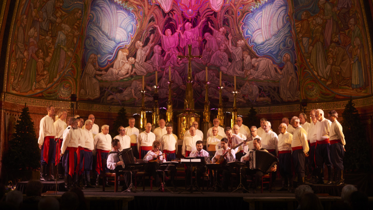 Een mannenkoor in witte blouses en rode sjerpen zingt op een podium met muzikanten ervoor, in een rijkelijk versierde kerk met muurmozaïeken en gouden kandelaars.