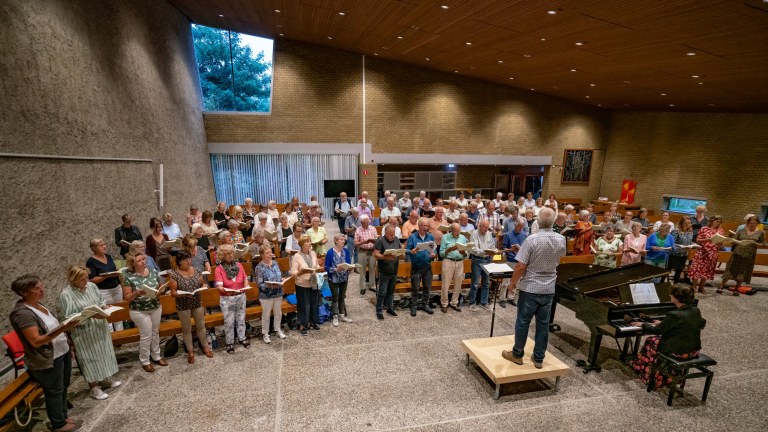 Koor repetitie in een kerkzaal met dirigent en pianist.