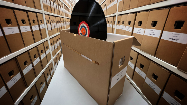 Kartonnen archiefdozen in lange rijen met een vinylplaat in een geopende doos.