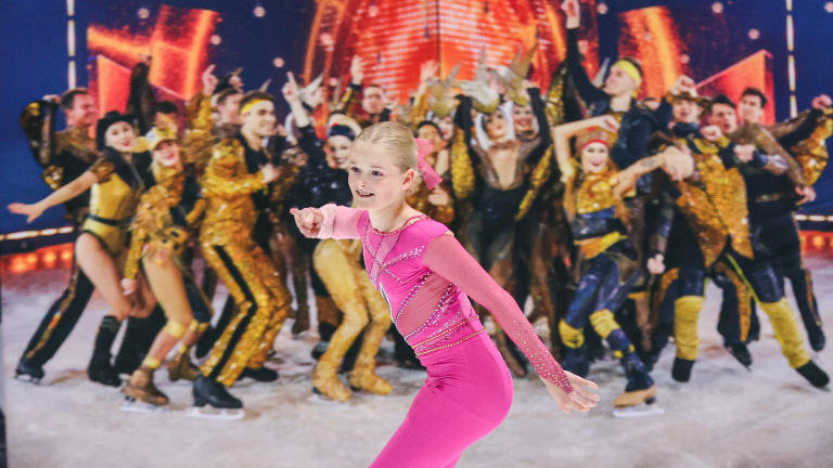 Schaatsster gekleed in roze met een groep mensen in gouden kostuums op de achtergrond tijdens een ijsvoorstelling.