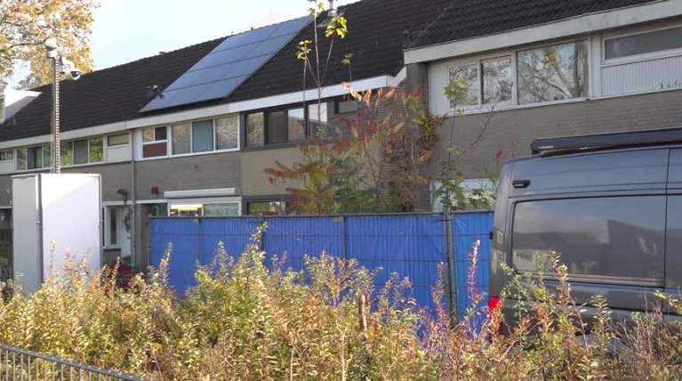 Rijtjeshuizen met zonnepanelen op het dak, omgeven door een blauwe schutting en begroeiing.