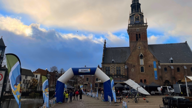 Een startboog met vlaggen op een plein bij een historisch gebouw met klokkentoren in Alkmaar.