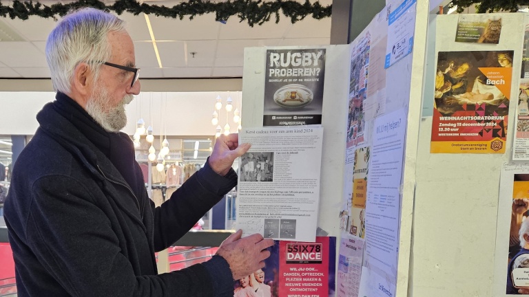 Een man met een grijze baard leest een flyer van een mededelingenbord in een winkelcentrum.