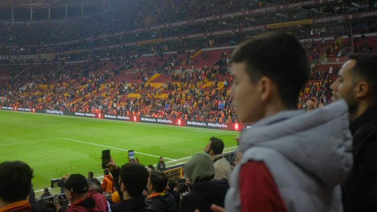 Jongen en man kijken naar voetbalwedstrijd in stadion vol met supporters, sommigen fotograferen.