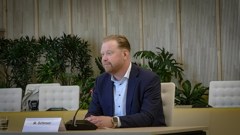 Man in pak zit aan tafel met microfoon; plant op de achtergrond.