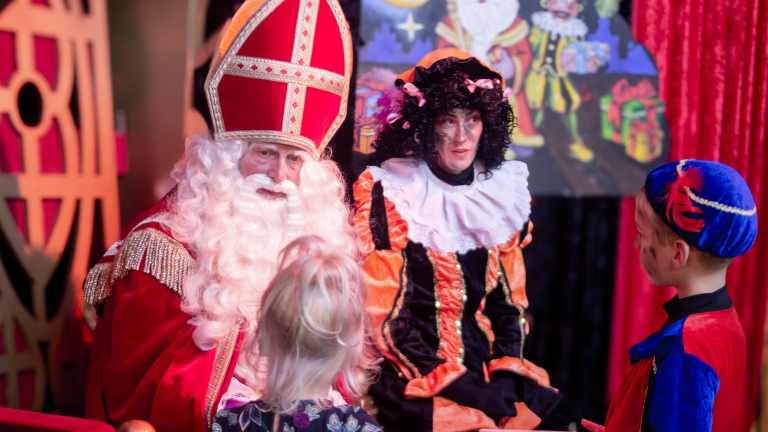 Sinterklaas en Zwarte Piet zitten op stoelen en praten met twee verklede kinderen.