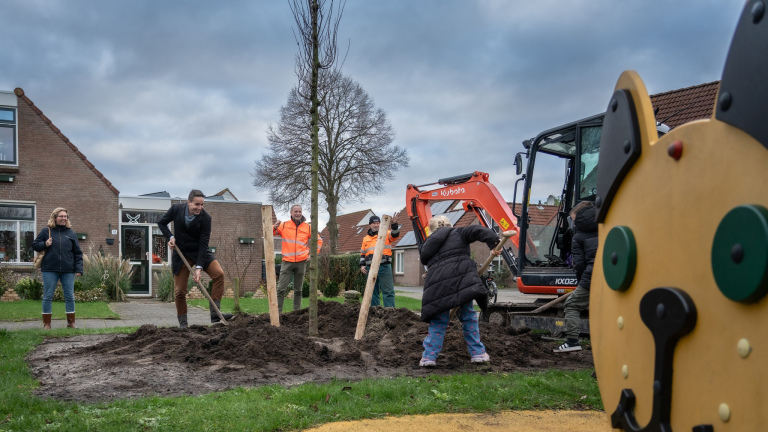 Mensen planten een boom in een woonwijk, met een graafmachine op de achtergrond.