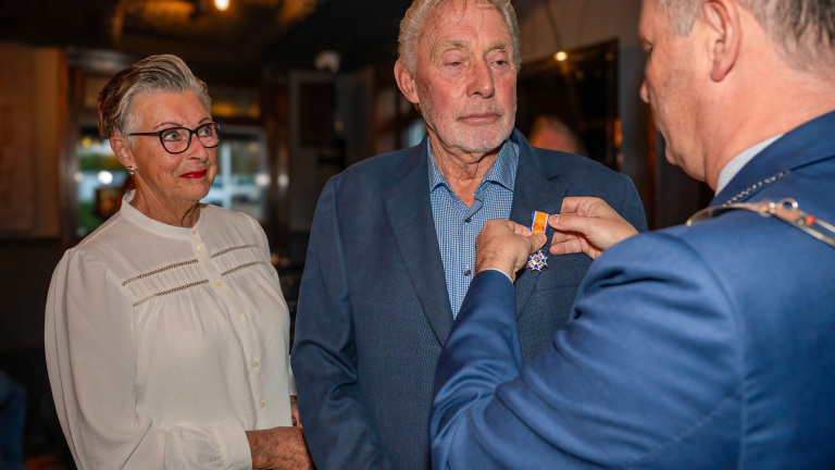 Een man ontvangt een Koninklijke onderscheiding, terwijl een vrouw en andere man toekijken.