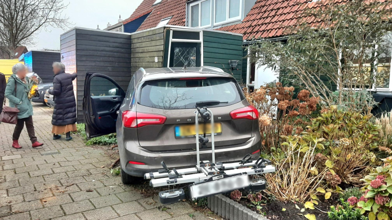 Auto geparkeerd op trottoir naast woning, met fietsendrager aan de achterkant; twee mensen lopen voorbij.