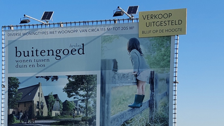 Reclamebord voor "Buitengoed Heiloo," met aankondiging van uitgesteld verkoop van woningen, tegen een blauwe lucht.