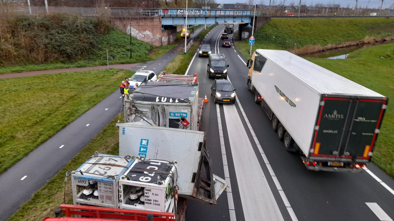 Vrachtwagen met aanhanger geblokkeerd op smalle weg, andere voertuigen passeren; mensen in veiligheidsvesten ernaast, brug op achtergrond.