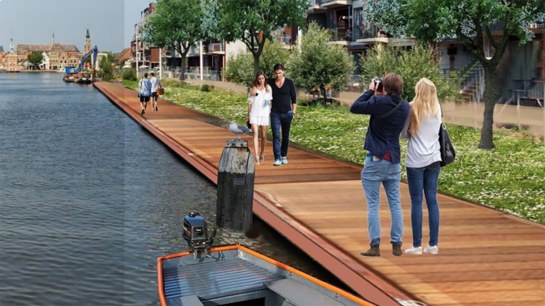 Een wandelpad langs het water met mensen die wandelen en fotograferen, omgeven door groen en gebouwen aan de overkant.