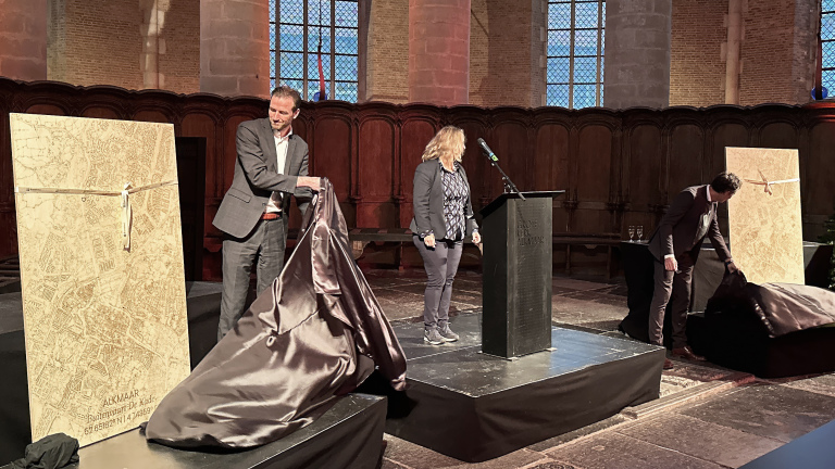 Twee mannen onthullen grote, gedetailleerde kaarten in een kerk terwijl een vrouw bij een spreekpodium staat.