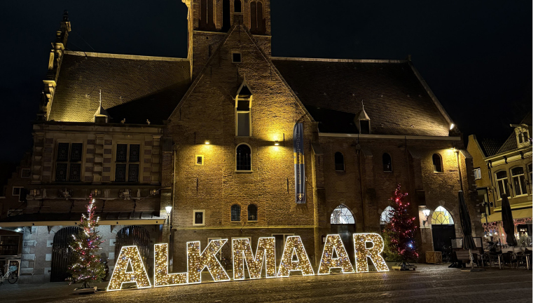 Verlicht gebouw met grote verlichte letters "ALKMAAR" en kerstbomen ervoor, 's nachts.