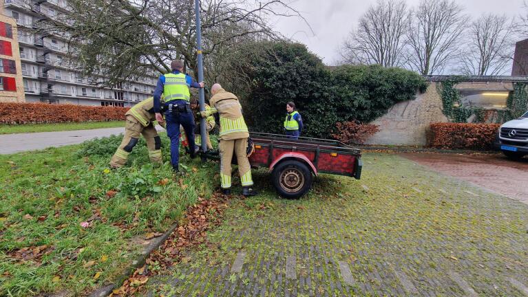 Enkele brandweermannen en handhavers werken naast een aanhanger bij een lantaarnpaal op een grasveld.