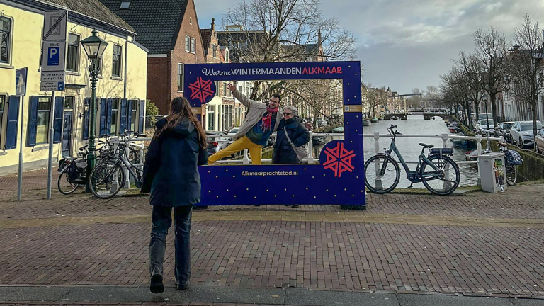 Een vrouw kijkt naar een fotokader met de tekst "Warme Wintermaanden Alkmaar" waarin twee mensen poseren; een kanaal en gebouwen op de achtergrond.