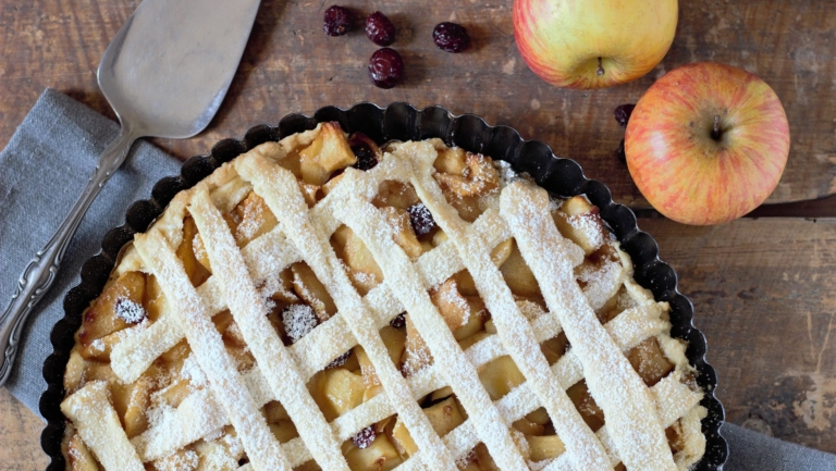 Appeltaart met een rasterpatroon van deeg, bestrooid met poedersuiker, op een houten tafel, naast twee appels en enkele gedroogde veenbessen.