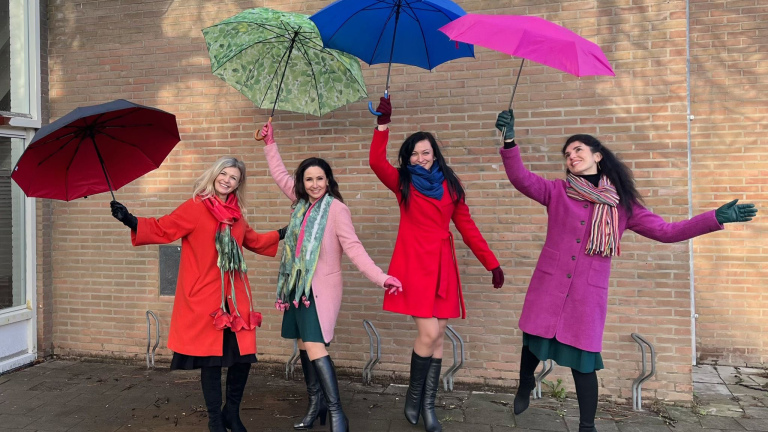 Vier vrouwen met kleurrijke jassen en paraplu's poseren vrolijk voor een bakstenen muur.