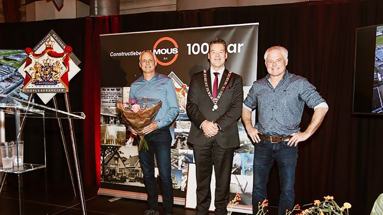 Twee mannen poseren met een burgemeester tijdens een jubileumviering van Constructiebedrijf Mous 100 jaar, naast een schild en banners.