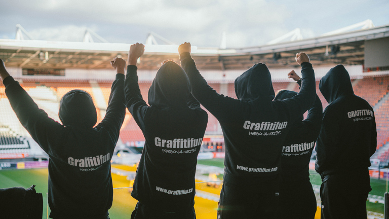 Vijf mensen in zwarte hoodies met "Graffitifun" erop priemen hun vuisten in de lucht in een stadion.